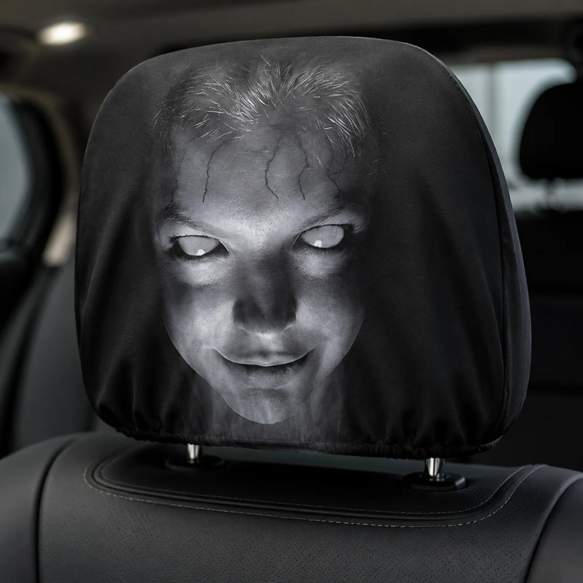 A car headrest cover featuring a creepy, horror-style printed face with pale glowing eyes and cracked skin, shown fitted over a black car seat headrest. 
Horror face cover installed over a black car headrest