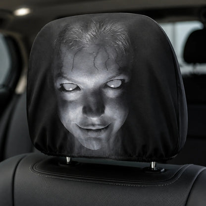 A car headrest cover featuring a creepy, horror-style printed face with pale glowing eyes and cracked skin, shown fitted over a black car seat headrest. 
Horror face cover installed over a black car headrest